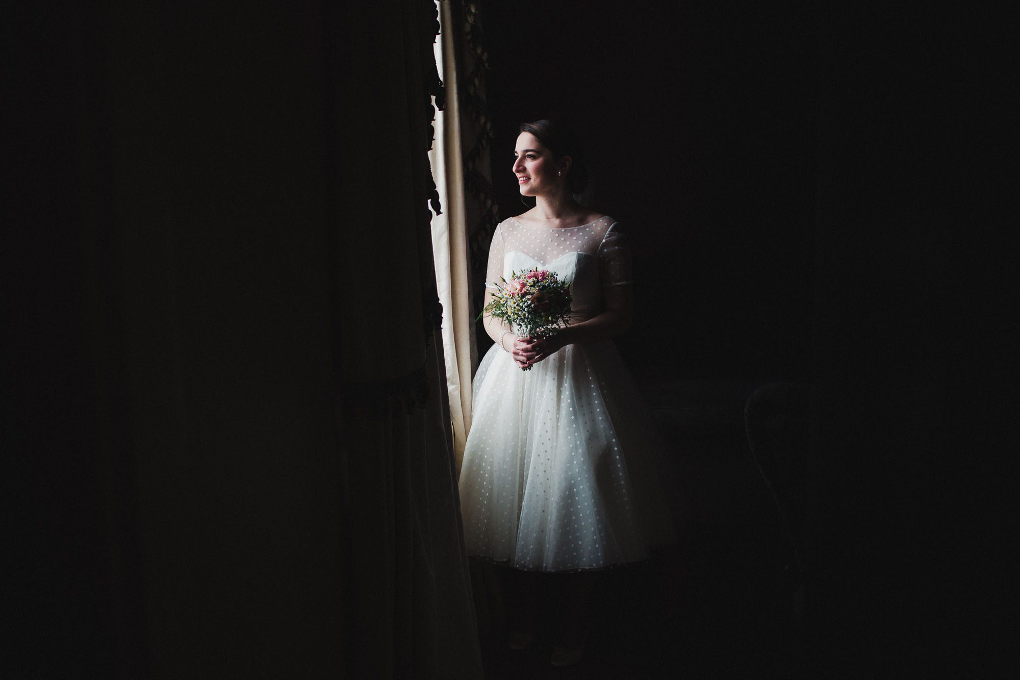 Bride standing bathed in window light. Wearing a spotty 50s style dress