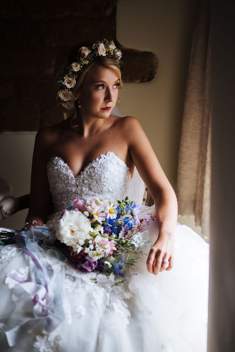 window light on bride as she sits next to window in her dress. Bride has bouquet on lap and is wearing a flower crown