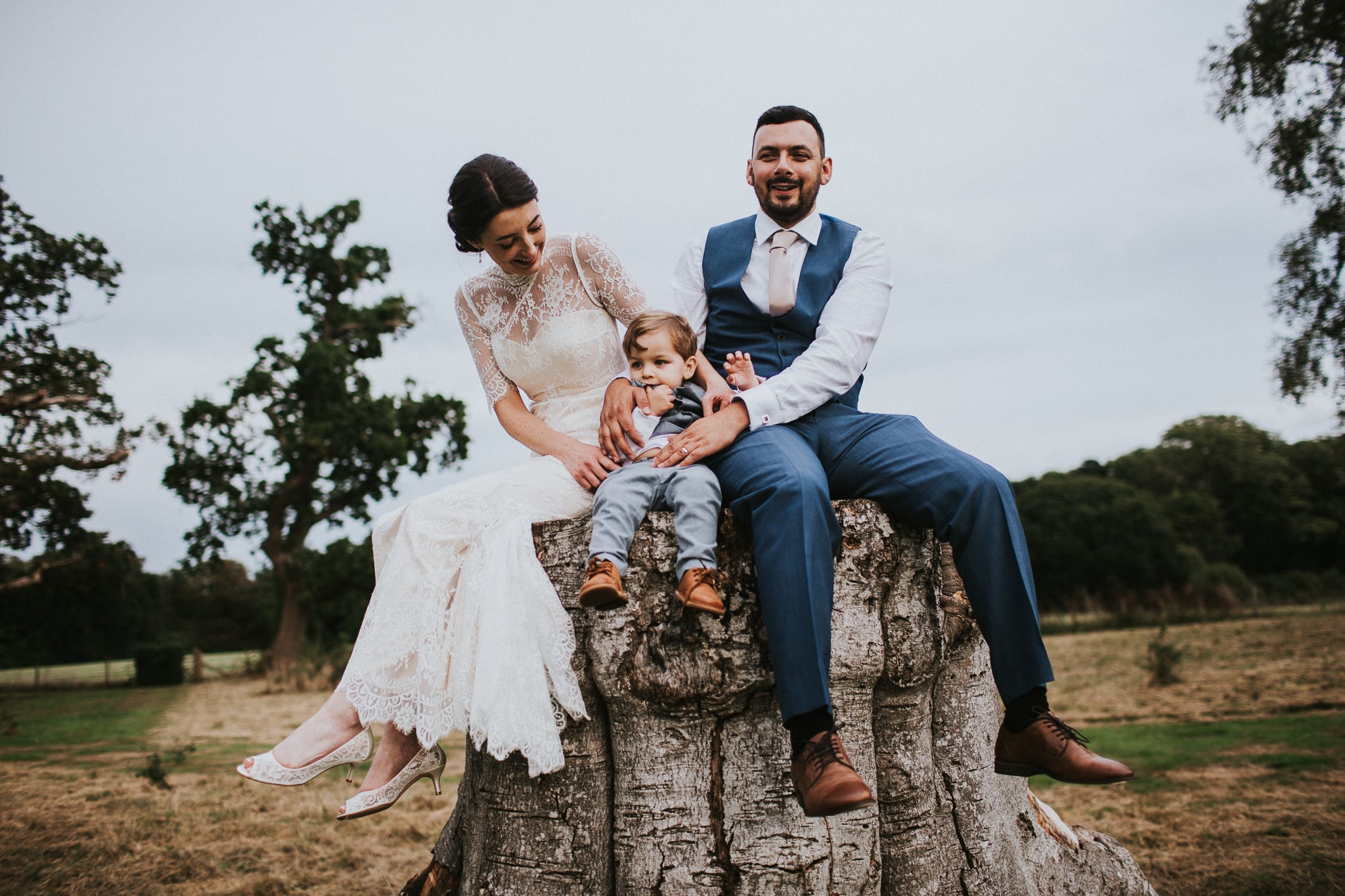 bride and groom and their 2 year old son sitting on a tree stump