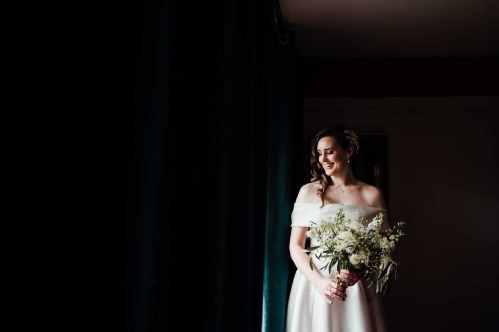 Bridal portrait - Louise holding bouquet looking out of the window. High contrast light.