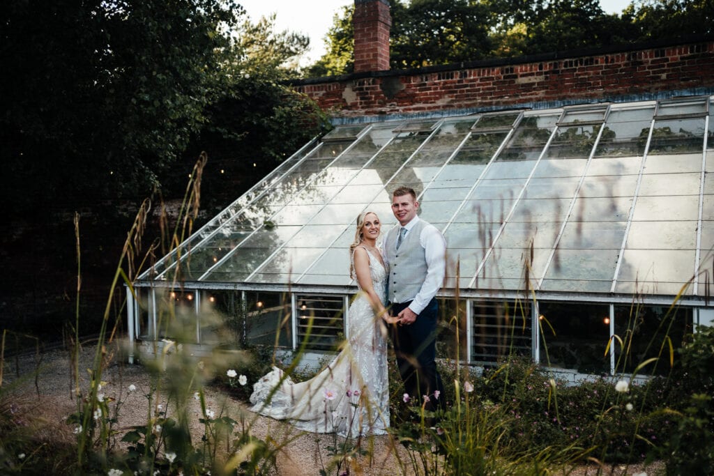 Bride and groom standing together in front of the greenhouse at The Walled Garden at Beeston Fields