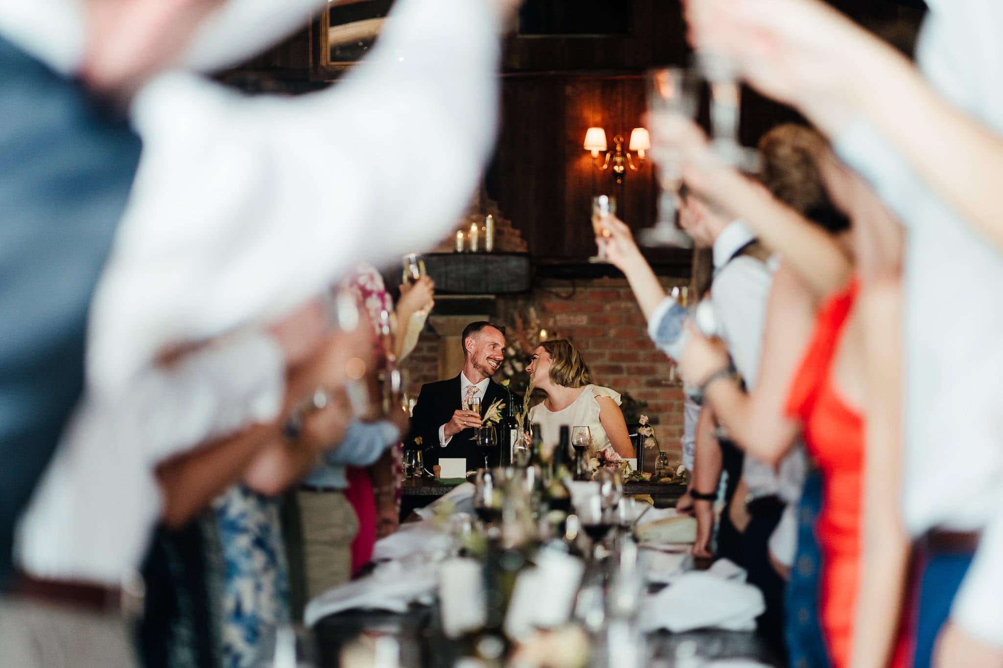 Bride and groom sit at end of table looking at each other whilst everyone is standing cheering their glasses, framing the couple along the long tables.